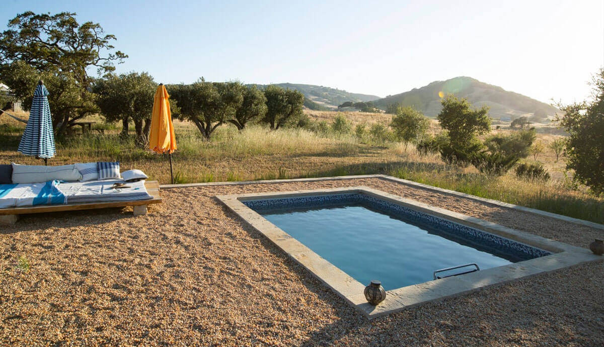 Endless Pools EP510 Modular Pool installed flush with a concrete edge in a rural open field setting, surrounded by golden grass and rolling hills in the background. A sun lounger with blue and white cushions sits nearby under striped and solid-coloured umbrellas, creating a relaxing, resort-like atmosphere.