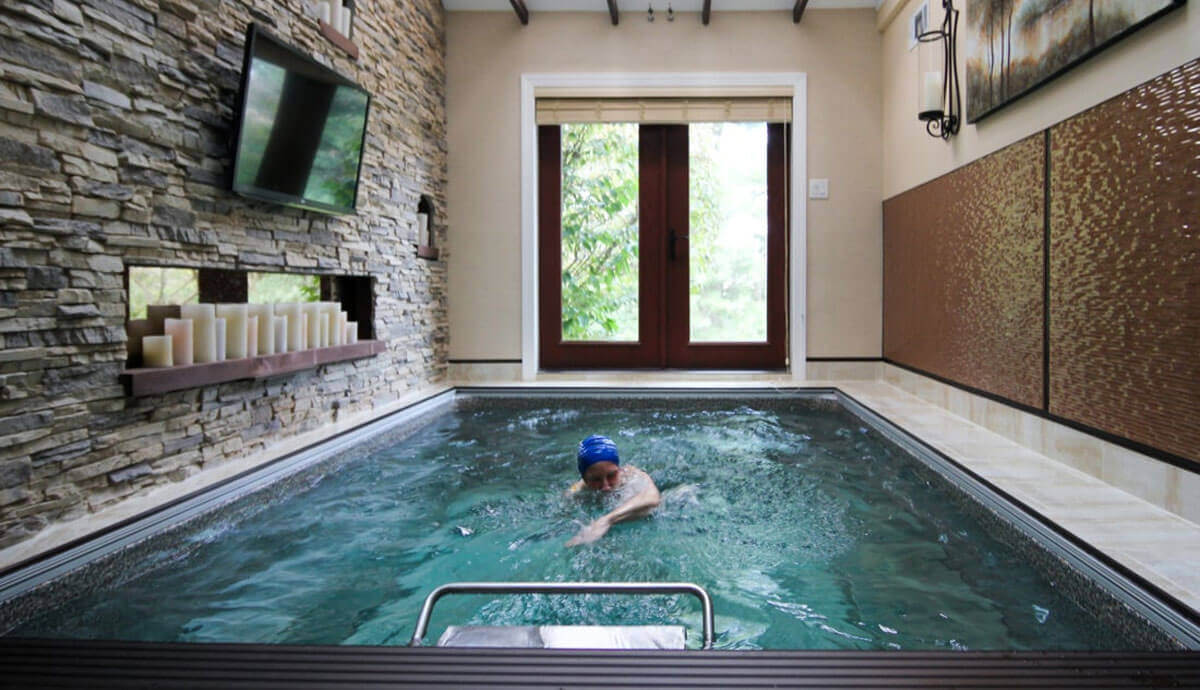 Indoor Endless Pools EP510 Modular Pool installation, set within a contemporary space featuring a stacked stone accent wall with built-in candle shelves and a mounted flat-screen television. A swimmer in a blue swim cap performs a freestyle stroke against the Hydrodrive™ current, while sunlight filters through double glass doors ahead.