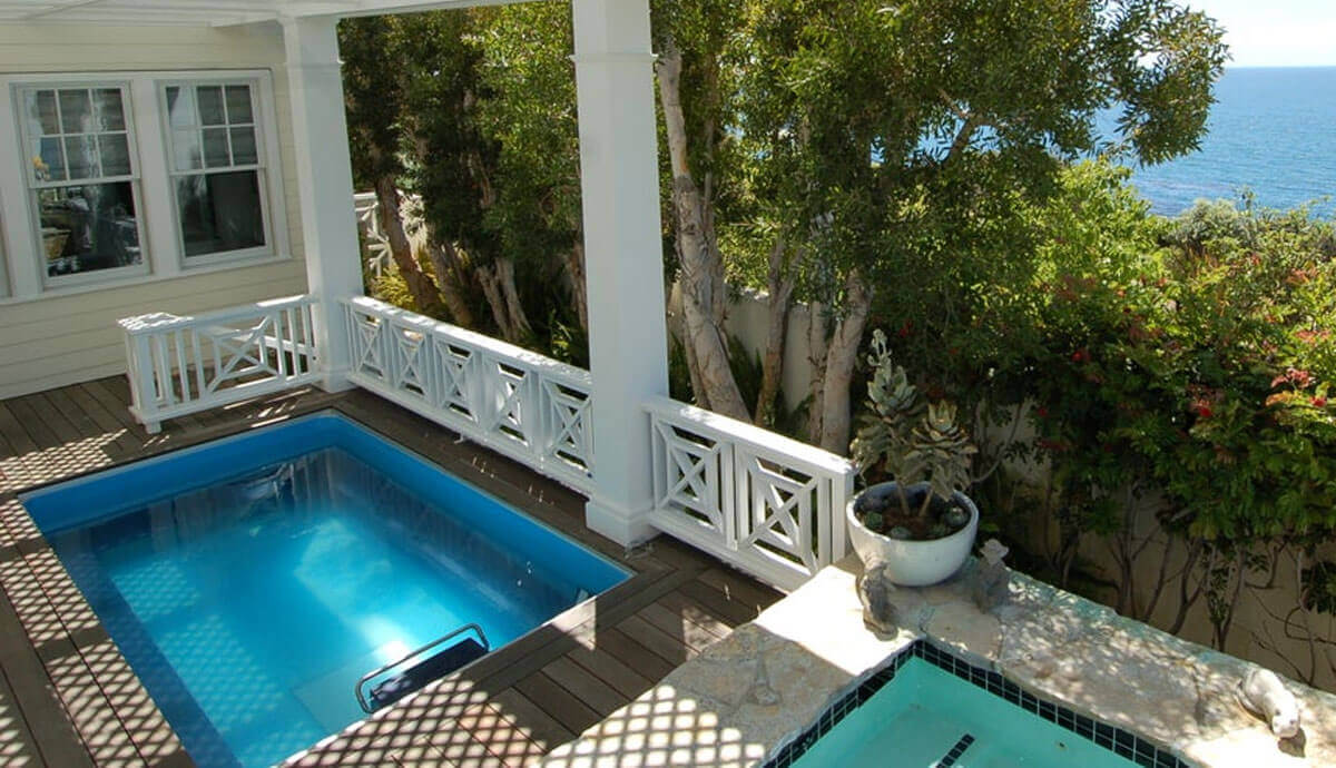 Endless Pools EP510 Modular Pool installed on a covered coastal balcony with a view of the ocean beyond. The bright blue water contrasts with the white railing and columns, while mature trees and flowering plants provide privacy. The pool sits adjacent to a connected raised spa, separated by stone coping.