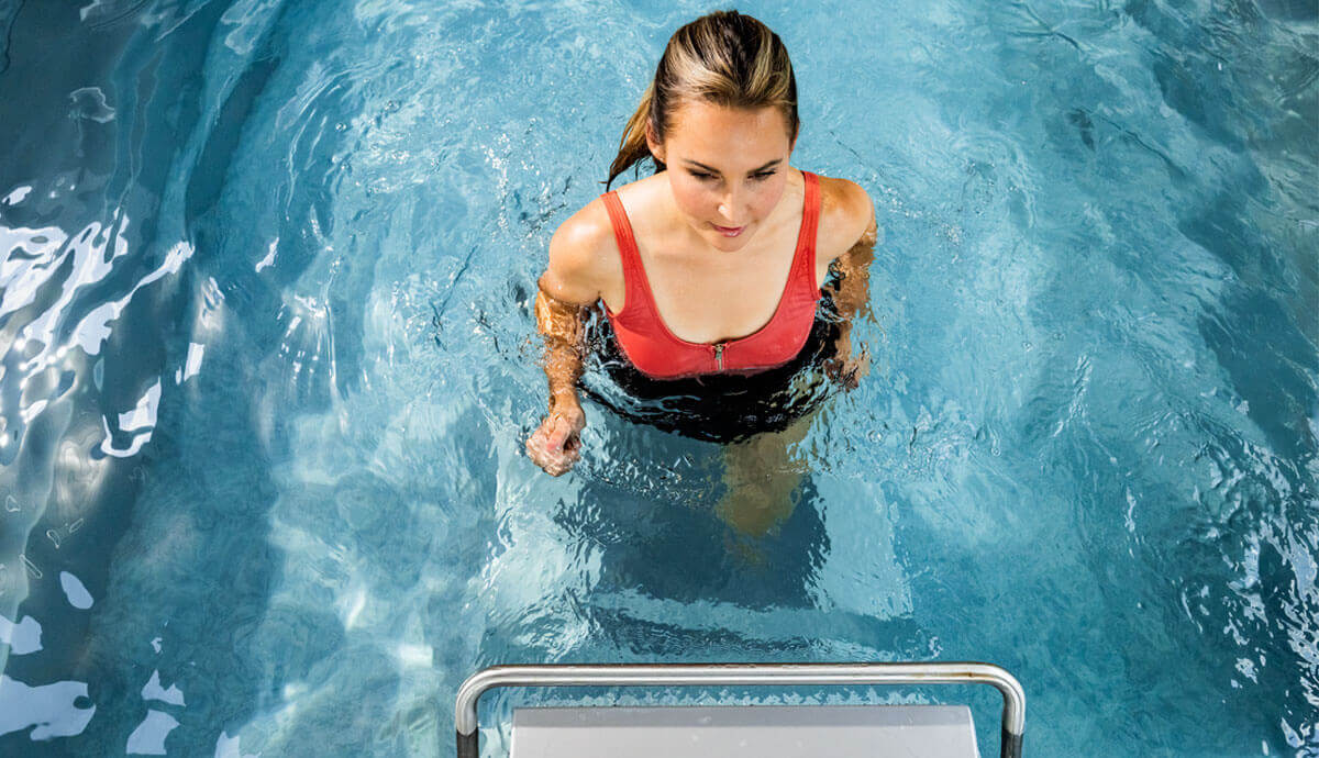 A woman wearing a bright red zip-front swimsuit enters the Endless Pools EP515 Plus Modular Pool using a stainless-steel ladder, with water rippling around her waist. The interior of the pool is lined with textured grey walls, and the bright sunlight creates shimmering reflections on the water surface.