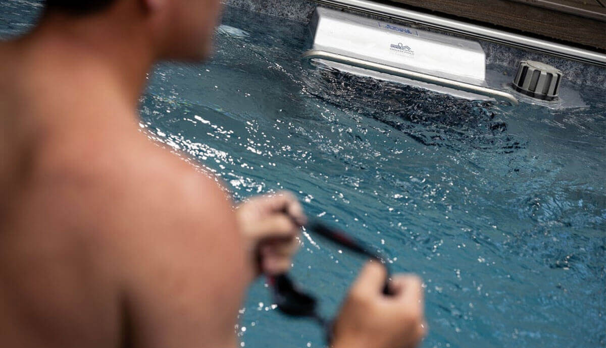Close-up view of the Hydrodrive Plus swim current unit inside the Endless Pools EP515 Plus Modular Pool, producing a powerful, smooth water flow. The silver housing with the Endless Pools logo is mounted centrally on the pool wall, next to a large grey flow adjustment dial. A swimmer’s blurred figure in the foreground prepares to train while holding swim goggles.