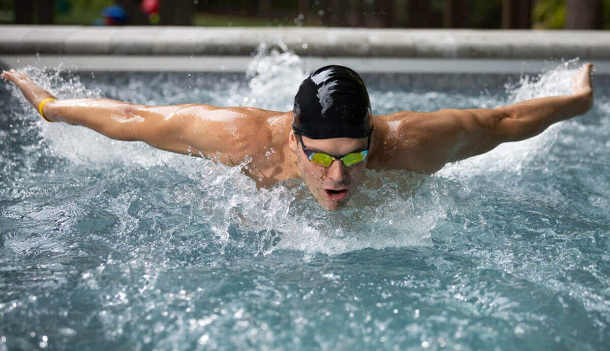 A male swimmer wearing a black swim cap and reflective goggles performs the butterfly stroke against the Hydrodrive Plus swim current in the Endless Pools EP515 Plus Modular Pool. Water splashes dynamically around his extended arms, with the textured grey pool wall visible in the background.
