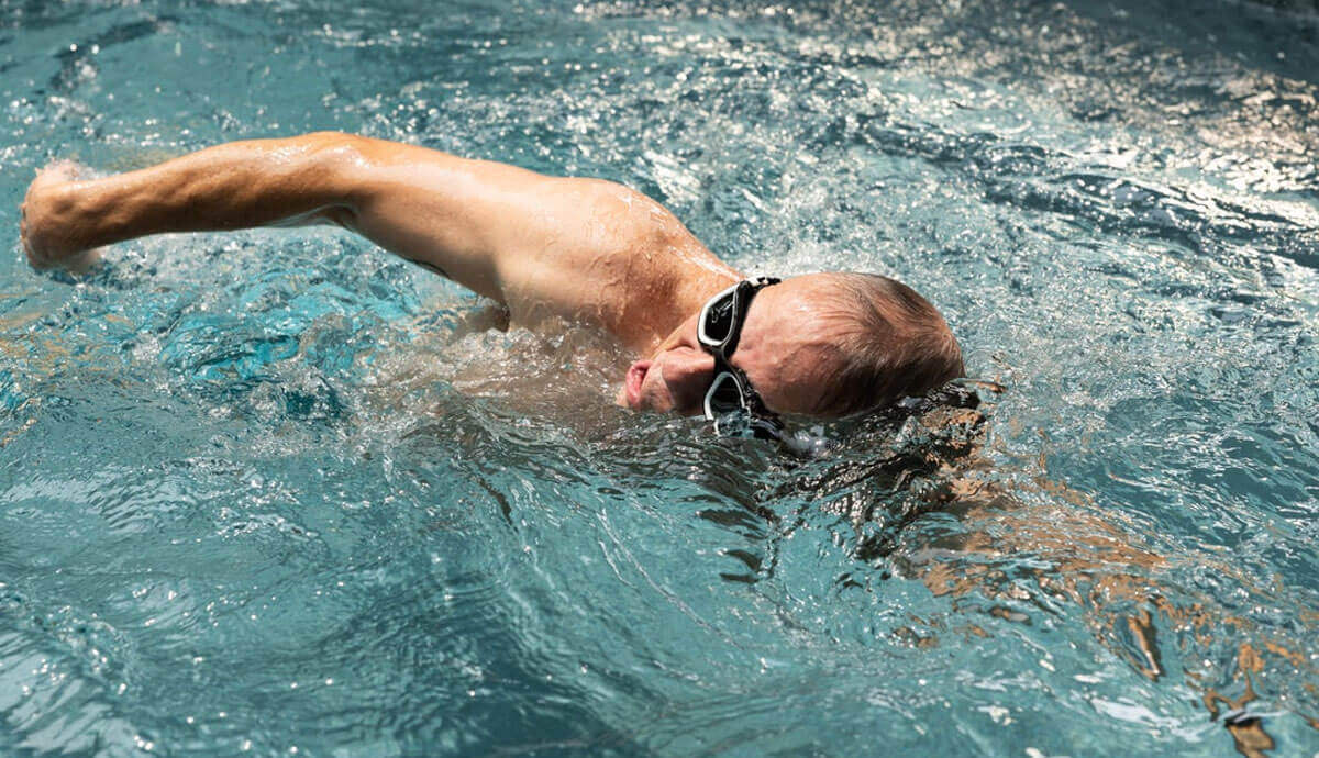 A male swimmer wearing black goggles and light blue swim shorts performs the freestyle stroke against the Hydrodrive Plus swim current in the Endless Pools EP515 Plus Modular Pool. Water turbulence surrounds his extended arm, with sunlight creating rippling highlights across the surface.