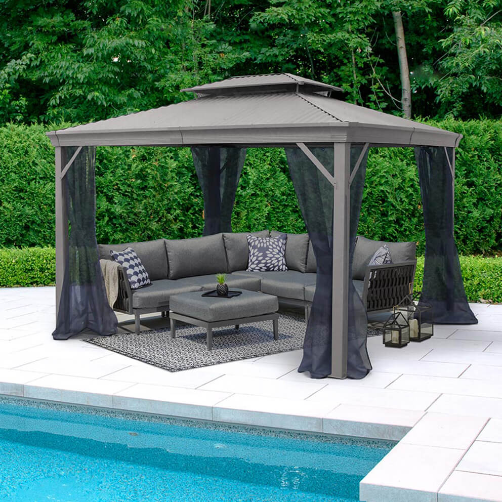 Outdoor view of the Maestrale Gazebo in a light grey finish, standing beside a swimming pool on a stone patio. The gazebo has a pitched Galvalume® metal roof with a raised vented section at the top for airflow, supported by four sturdy square aluminium posts with decorative base covers. Sheer black mosquito net curtains hang loosely from the roof frame, gathered neatly at the posts. Under the gazebo sits a full grey sectional outdoor sofa with patterned accent pillows and a central ottoman on a rectangular rug. A pair of lanterns are placed by the corner of the gazebo, and lush green hedges provide a natural privacy backdrop.