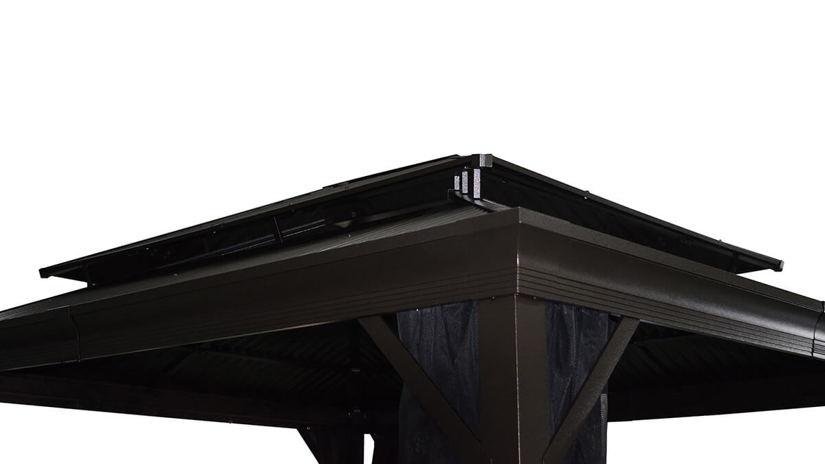 Black Maestrale Gazebo installed on a raised wooden deck surrounded by trees and greenery. The gazebo has a sleek pitched Galvalume® metal roof with a vented second tier, designed to allow airflow and stability. Four square aluminium posts with strong decorative bases support the frame, with sheer black mesh mosquito curtains tied neatly around each post. The surrounding deck features chevron-patterned wooden planks, a potted plant in the corner, and a backdrop of tall evergreen trees, creating a natural and private retreat.