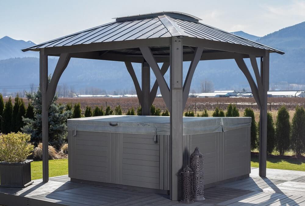 A Napa Gazebo with a pitched dark gray roof and open sides, installed over a square hot tub. The gazebo stands on a raised deck surrounded by grass and landscaped shrubs. Two decorative lanterns are placed beside the hot tub, and the scene overlooks distant mountains under a clear sky.