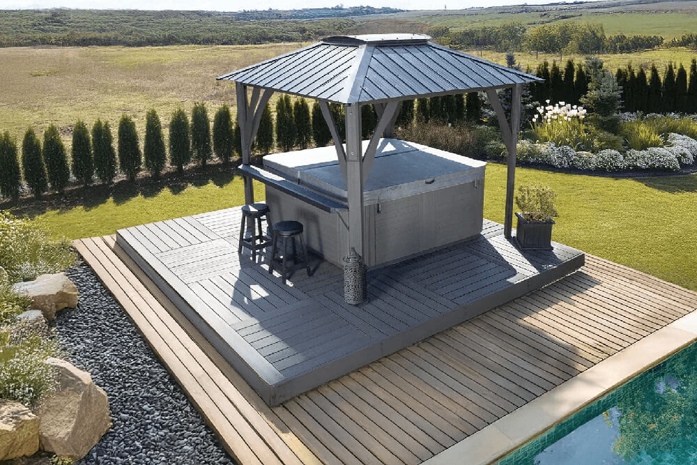 A top angled view of the Napa Gazebo covering a spa, set on a gray wooden deck with light wood border trim next to a swimming pool. Two black bar stools sit at one side of the hot tub under the gazebo. Decorative lanterns and potted plants accent the deck, while hedges and flowering shrubs surround the lawn in the background.