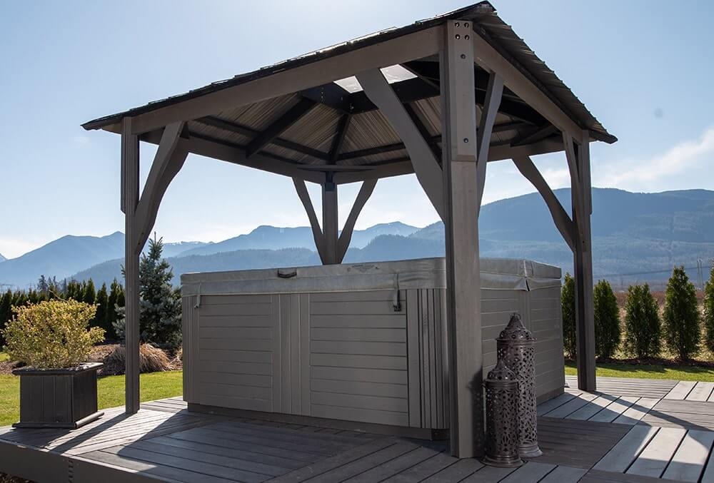 Close perspective of the Napa Gazebo with its roof supported by four wooden-style posts, installed above a rectangular hot tub. The spa has light gray siding and sits on a raised deck. Two ornate lanterns stand on the deck beside the tub. The background shows neatly trimmed hedges and distant mountain ranges.