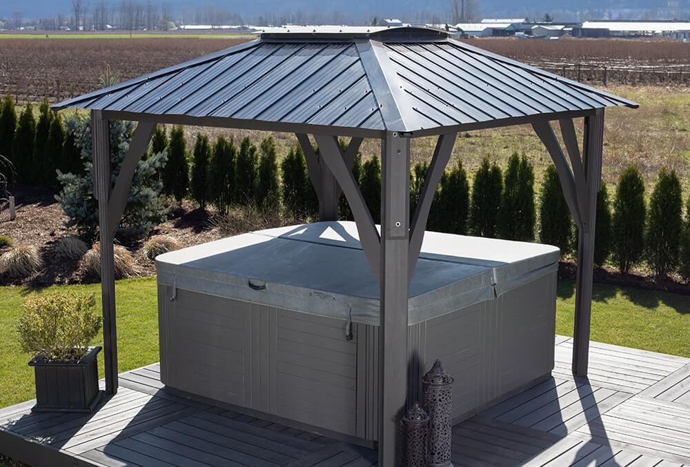 Side view of the Napa Gazebo with its ribbed metal roof sloping downward, covering a rectangular spa with a light gray exterior. Decorative lanterns are placed near one post, and the surrounding lawn features small shrubs and hedges, with farmlands visible in the distance.