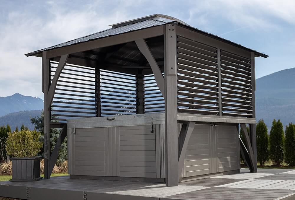 Rear three-quarter view of the Napa Gazebo, showing its slatted adjustable louver wall panels partially closed for shade and privacy. The structure sits above a hot tub with light gray siding, placed on a gray raised deck. Trees, hedges, and mountain ranges form the backdrop under a lightly clouded sky.