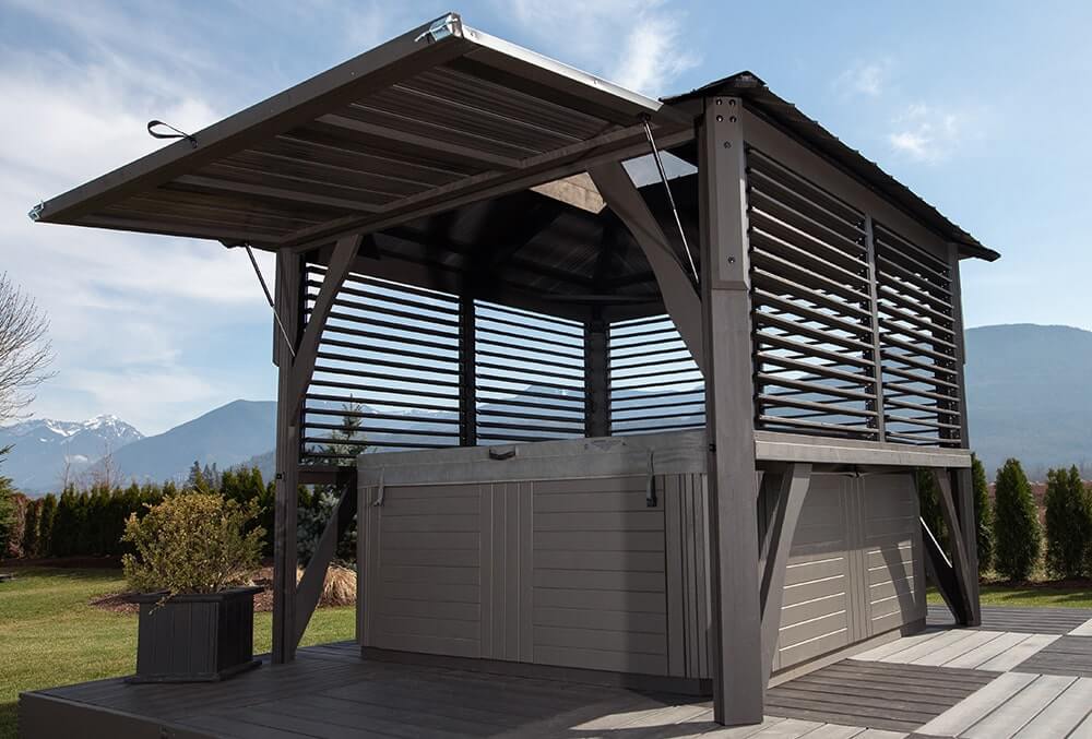 Angled side view of the Napa Gazebo, displaying its adjustable louver wall panels fully open on one side. The slats are supported by sturdy posts and a pitched ribbed roof above. A hot tub with gray siding sits inside, surrounded by a raised gray deck and landscaped lawn with mountain scenery in the background.