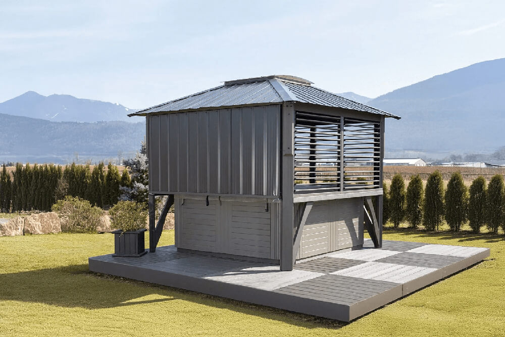 Rear perspective of the Napa Gazebo with both side walls fully covered by horizontal adjustable louvers and the back wall closed with solid gray panels. The gazebo covers a spa and sits on a raised wooden deck. Evergreen hedges and distant mountains fill the background.
