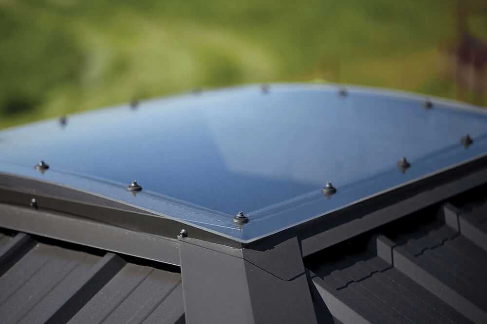 Close-up of the Napa Gazebo skylight dome at the roof peak. The skylight is a translucent panel bordered by black framing and secured with evenly spaced fasteners. The roof itself is ribbed dark gray metal, with blurred green landscape visible in the background.