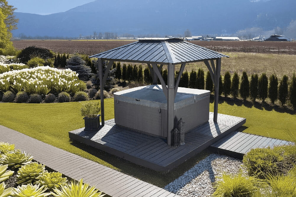 Wide garden view of the Napa Gazebo installed over a spa on a raised deck platform. The gazebo roof has a pronounced skylight dome at the centre, supported by four sturdy posts with diagonal braces. Surrounded by lush landscaping, white flowering plants, and a row of tall hedges, the gazebo forms a tranquil outdoor retreat with mountain scenery in the distance.