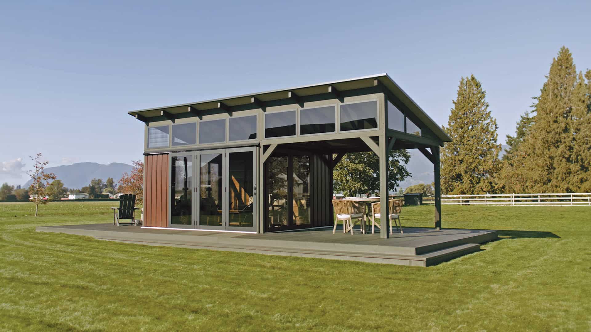 Outdoor view of the Oslo Gazebo positioned on a grassy field, showcasing its asymmetrical roofline sloping down to the right. The left side is enclosed with tall glass doors and wood-paneled walls, while the right side is open with a dining set placed on the covered patio.