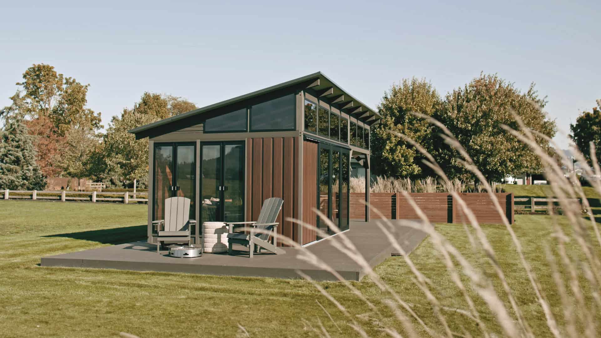 Rear corner perspective of the Oslo Gazebo, emphasizing its combination of large glass sliding doors, dark aluminum framing, and vertical reddish-brown wooden siding. Two lounge chairs are positioned outside, with ornamental grasses in the background.
