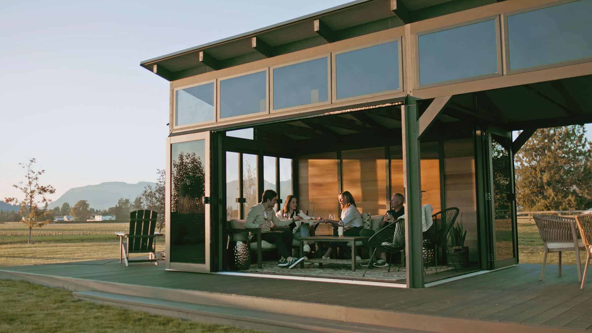 Close-up side view of the Oslo Gazebo with its bi-fold glass doors fully opened, revealing an indoor living space. Four people sit around a low coffee table, with cushioned chairs and a rug inside, while an outdoor dining set is visible through the opening.