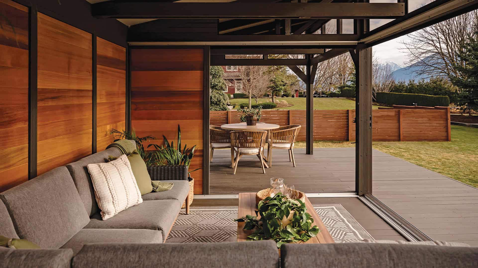 Interior view of the Oslo Gazebo looking outwards, showing a cozy living space with a gray sectional sofa, green accent pillows, a patterned area rug, and potted plants. The opposite side features an outdoor round dining table with wooden chairs, visible through the wide open sliding doors.