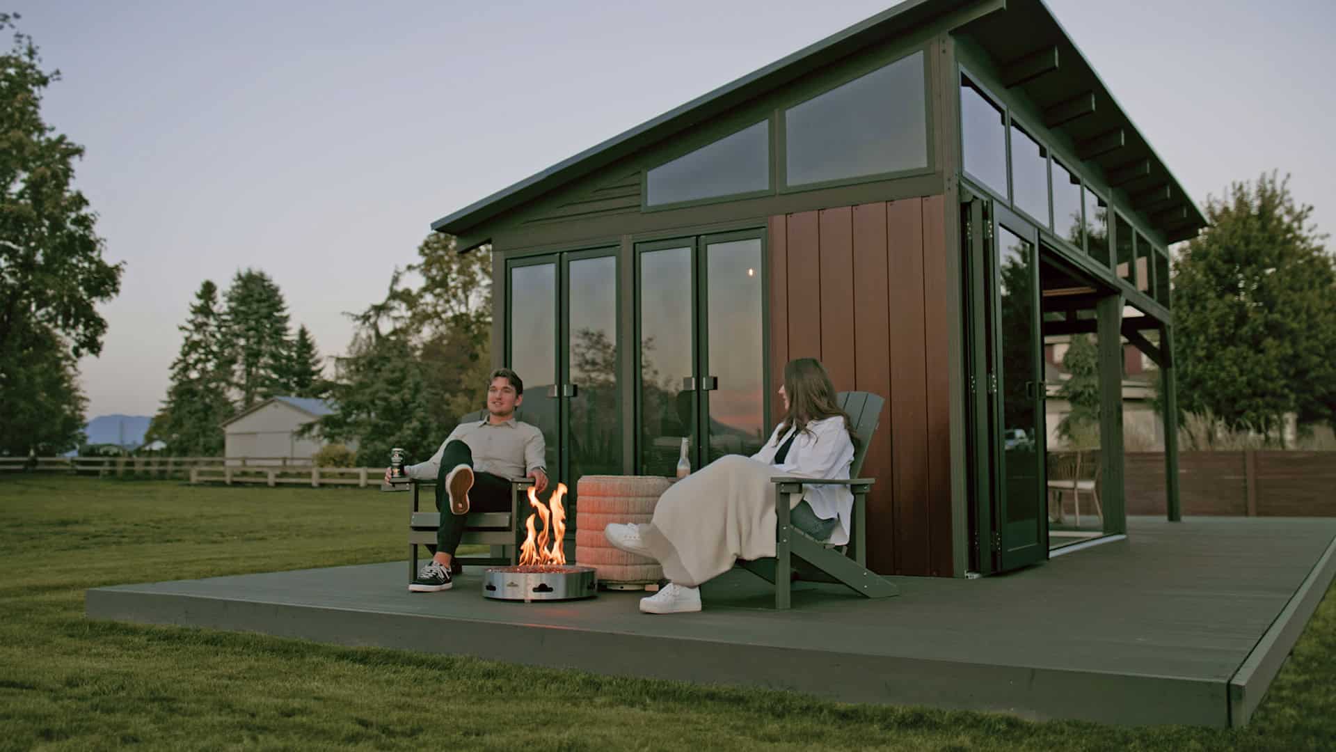 Evening scene of the Oslo Gazebo with a couple seated outside in Adirondack chairs around a firepit. The glass doors reflect the dimming sky, while warm light glows softly from the inside, highlighting the wooden siding and pitched roofline.