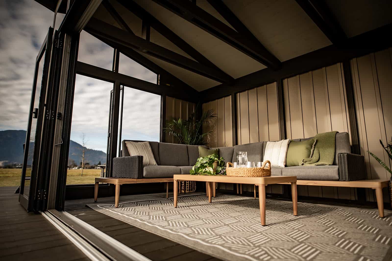 Interior corner perspective of the Oslo Gazebo, focusing on a modern sitting area. The space features a woven sectional sofa with green and cream pillows, a patterned area rug, and a wooden coffee table decorated with a tray, glassware, and greenery. Large folding glass doors stand open, framing the outdoor view and allowing natural light inside.