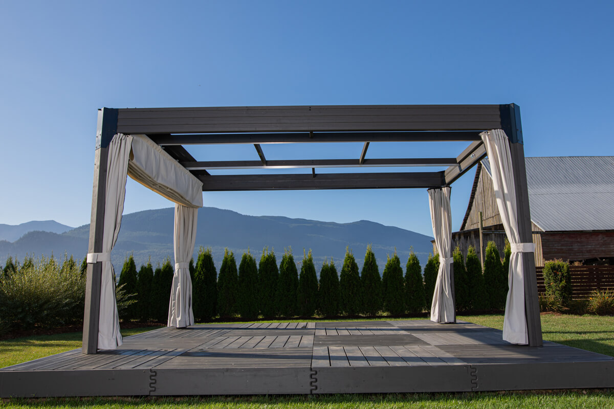 Open-roof Palmera Gazebo photographed outdoors with a mountain range in the background. The frame is constructed from sleek, dark grey posts and beams, with cream-coloured curtains neatly tied back at each corner. The top roof beams are uncovered, showing the open slatted support system for the retractable sunshade. The structure sits atop a raised grey platform deck, creating a modern outdoor space with clear, unobstructed sky above.