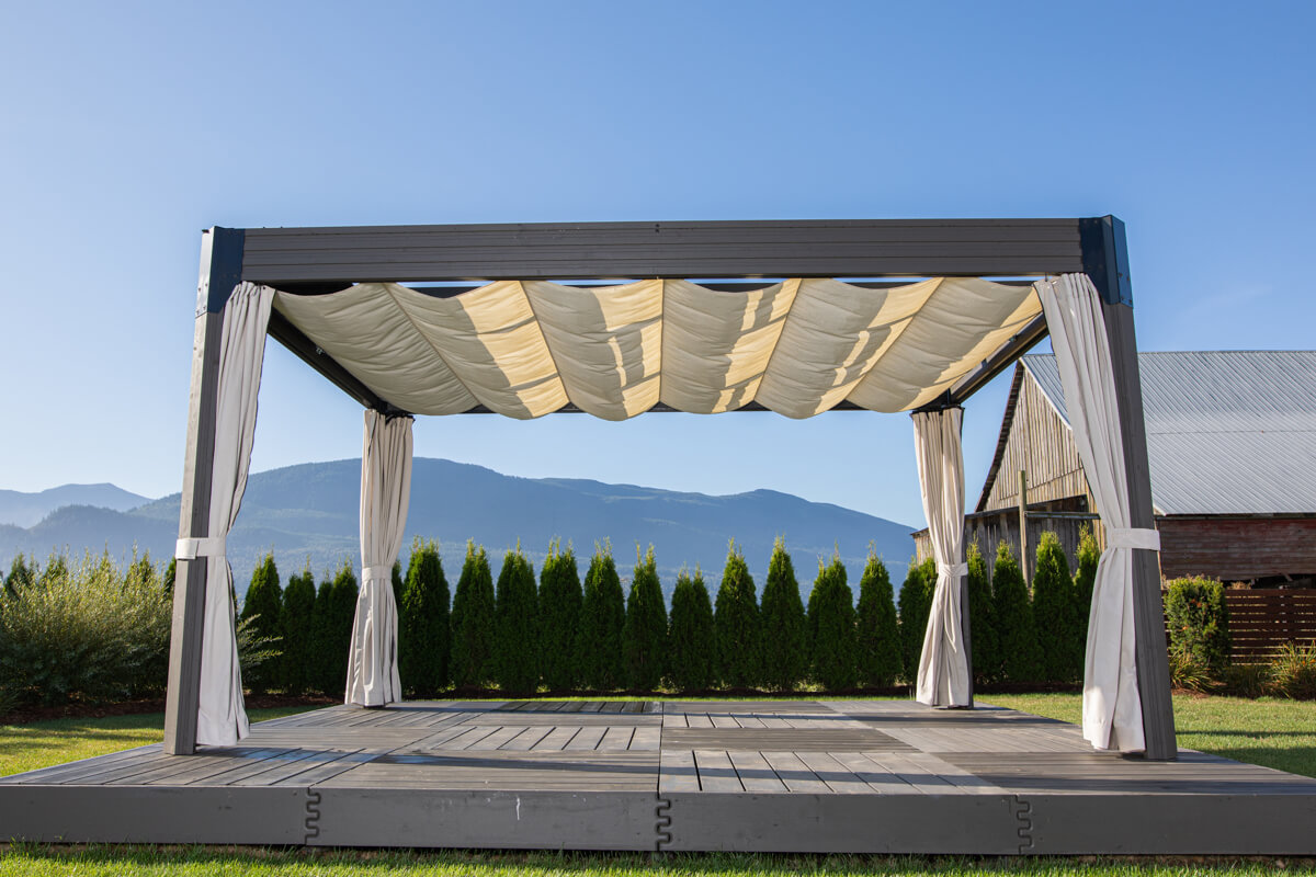 Palmera Gazebo with retractable cream-coloured canopy fully extended across the top, creating soft fabric folds that provide shade. The dark grey square posts support the modern rectangular frame, while all four curtains are tied back to the posts. The gazebo is set on a matching raised grey deck with tall evergreens behind, and the mountains visible in the distance under a clear blue sky.