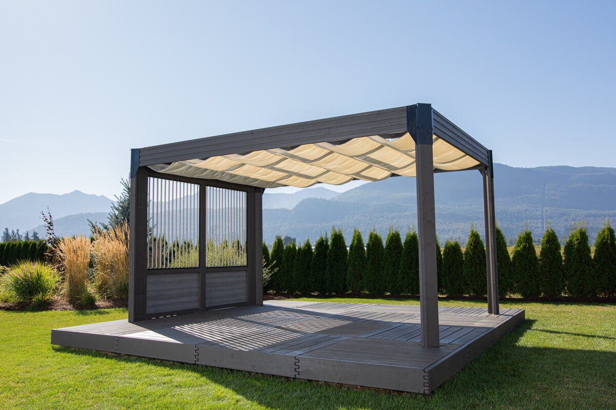 Side angle of the Palmera Gazebo showing the retractable sunshade fully deployed in soft cream-coloured waves across the roof. The structure is fitted with vertical slatted privacy panels on one side, framed in dark grey to match the posts and beams. The gazebo is positioned on a raised composite deck in a landscaped garden with ornamental grasses and hedges, with mountains in the background.