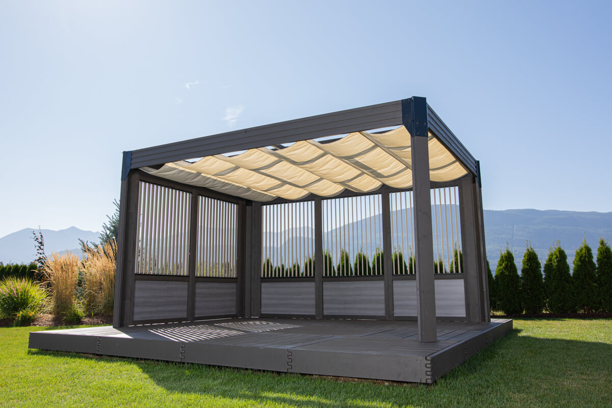 Angled view of the Palmera Gazebo with cream-coloured sunshade canopy deployed across the top. The gazebo includes vertical privacy slats on two sides, providing semi-enclosure while allowing light and air through. The dark grey modern frame sits on a raised grey deck, surrounded by neatly trimmed grass, tall hedges, and scenic mountain views in the background.