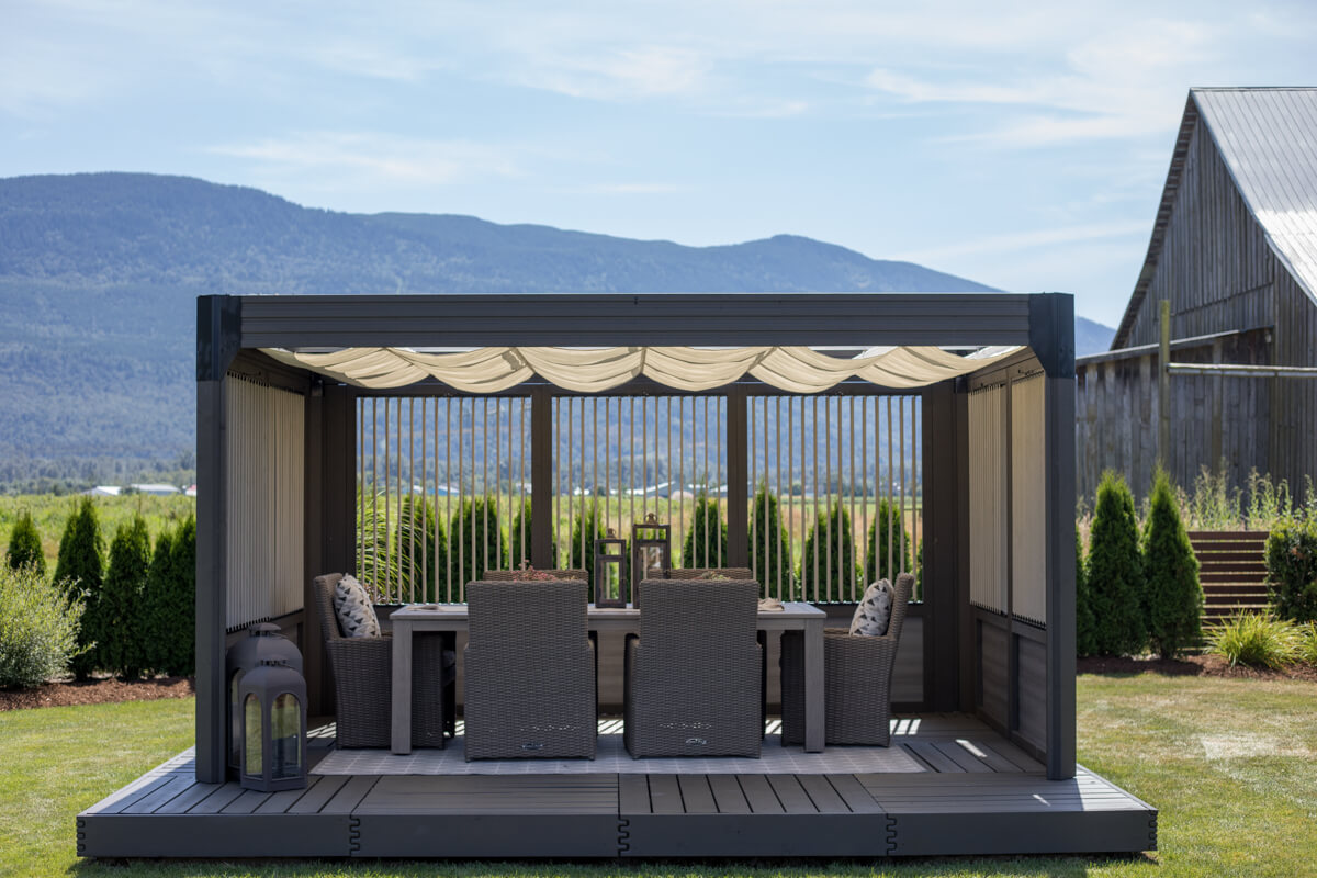 Palmera Gazebo staged as an outdoor dining space, with wicker-style armchairs arranged around a rectangular dining table placed inside. Decorative lanterns sit on the deck and tabletop, enhancing the cosy atmosphere. The cream-coloured canopy overhead filters the sunlight, while the vertical slatted privacy panels on three sides provide structure and shade. The gazebo rests on a raised grey deck against a scenic background of trees, hedges, and mountains.