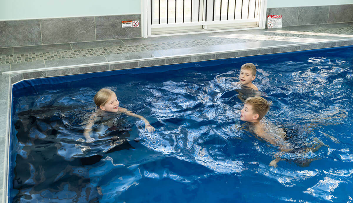 Three children playing inside the Endless Pools Plunge Max Therapy Pool filled with deep blue water indoors. The pool’s edge is lined with grey tile flooring and a decorative strip of small round mosaic tiles. A white safety gate is visible in the background. The children are mid-conversation, treading water and smiling, with ripples creating reflective patterns on the water surface.