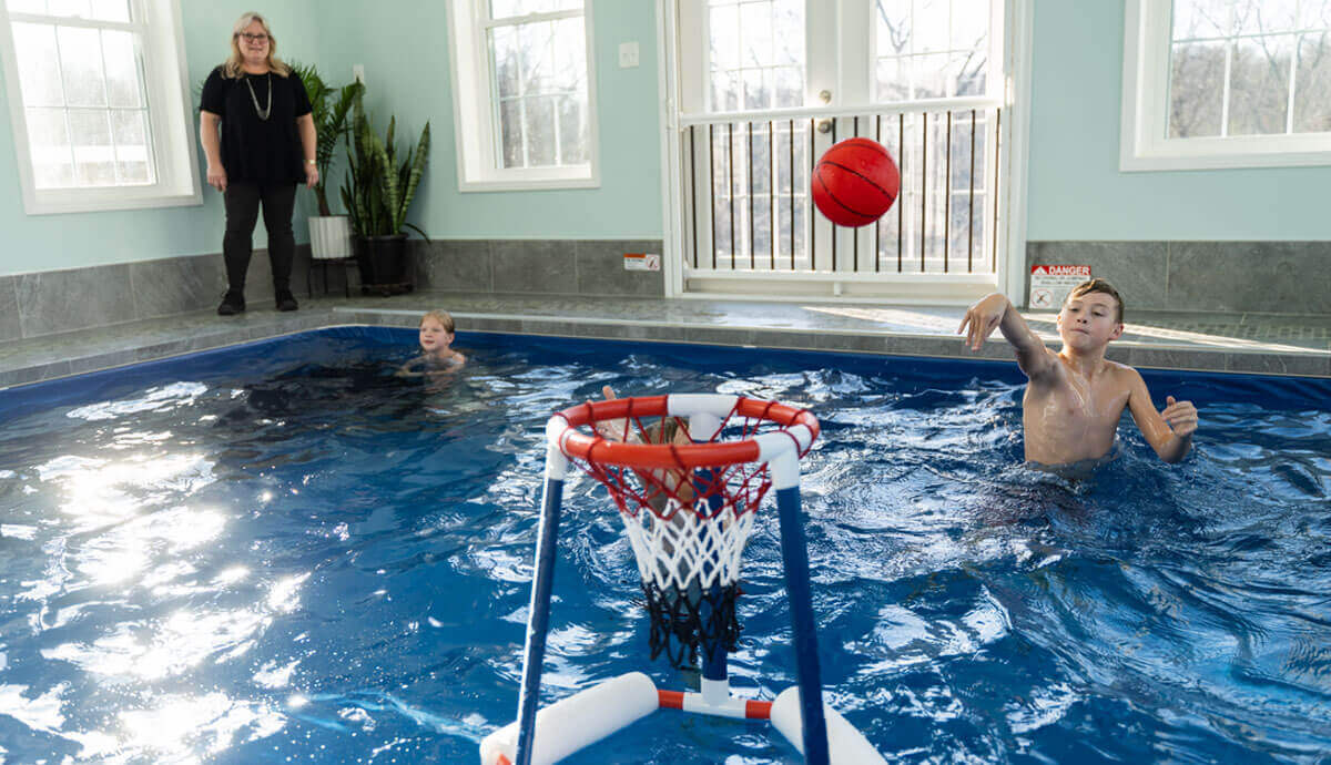 Indoor view of the Endless Pools Plunge Max Therapy Pool filled with water, where two boys play basketball while a woman stands nearby on the grey-tiled pool deck. The nearer boy is shooting a bright red basketball toward a floating basketball hoop with a white net, red rim, and blue base positioned in the water. A second boy is partially submerged in the background. Behind them is a white safety gate with glass double doors and two large windows allowing daylight into the space.