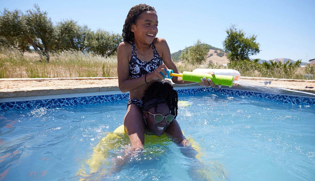 Two children playing in a bright outdoor in-ground pool. A girl wearing a black patterned bikini sits on the shoulders of another child, holding a green and white water blaster toy spraying a strong stream of water. The pool has a blue patterned liner and beige coping. The background features grassy fields, olive trees, and distant hills under a clear sky.
