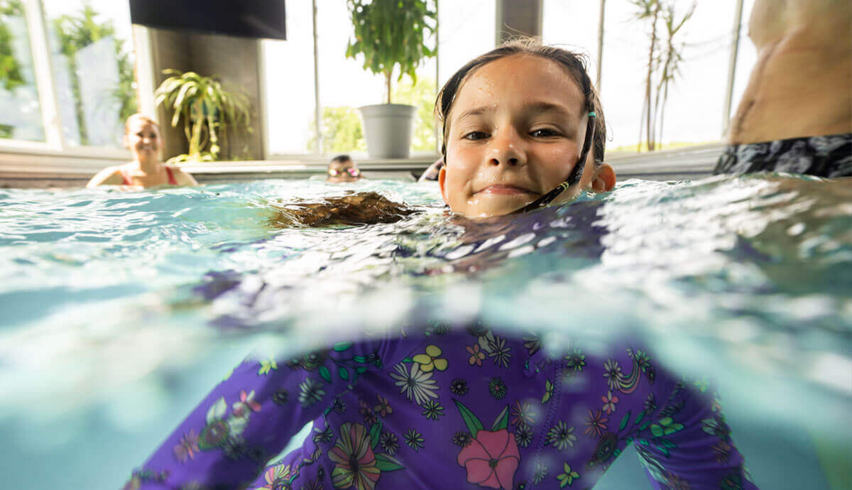 Close-up, partially underwater view of a smiling girl wearing a purple long-sleeved swim shirt decorated with colourful flowers, rainbows, and stars inside an indoor pool. The waterline cuts across the middle of the frame, with light reflecting off the rippled surface. In the background, an adult and another child are visible out of focus, along with large potted plants and windows letting in bright daylight.