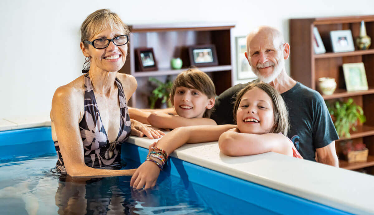 Family gathered around the Endless Pools Plunge Mini Therapy Pool indoors. A woman in a floral-patterned swimsuit and black glasses stands in the water smiling, while two children lean on the white coping edge smiling toward the camera. A man with white hair and a beard stands behind them. The interior water is a vibrant blue, and the background shows bookshelves with framed pictures and plants.