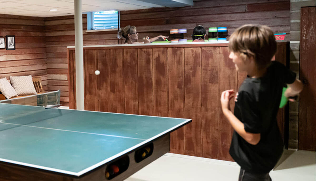 Basement recreation room with a ping pong table in the foreground, a boy in motion holding a green paddle, and the Endless Pools Plunge Mini Therapy Pool visible in the background. The pool is enclosed in wood-panelled exterior walls, with two people partially visible inside, wearing goggles and aquatic gear. Lighting is warm, and the wall panels have a reddish-brown wood grain.