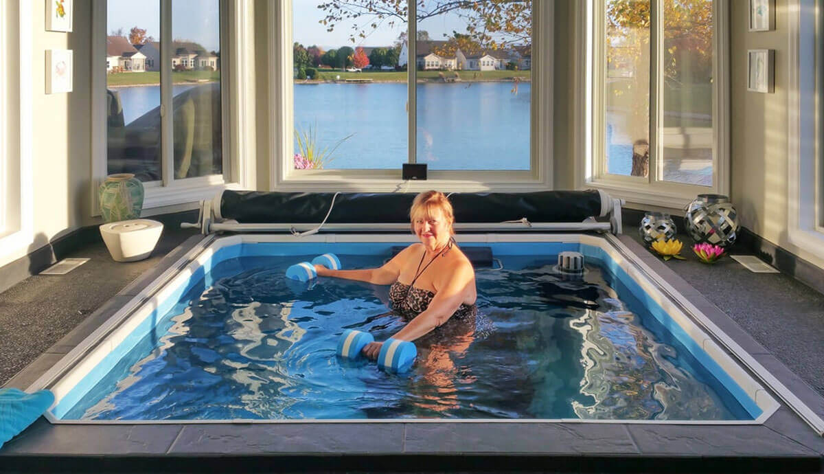 Indoor installation of the Endless Pools Plunge Prime Therapy Pool, surrounded by a black tiled floor and large panoramic windows overlooking a lake. A woman in a patterned swimsuit is performing water exercises using a pair of light blue foam aquatic dumbbells. The white slatted cover roller is retracted at the far side. The water is gently rippled, reflecting the bright daylight.