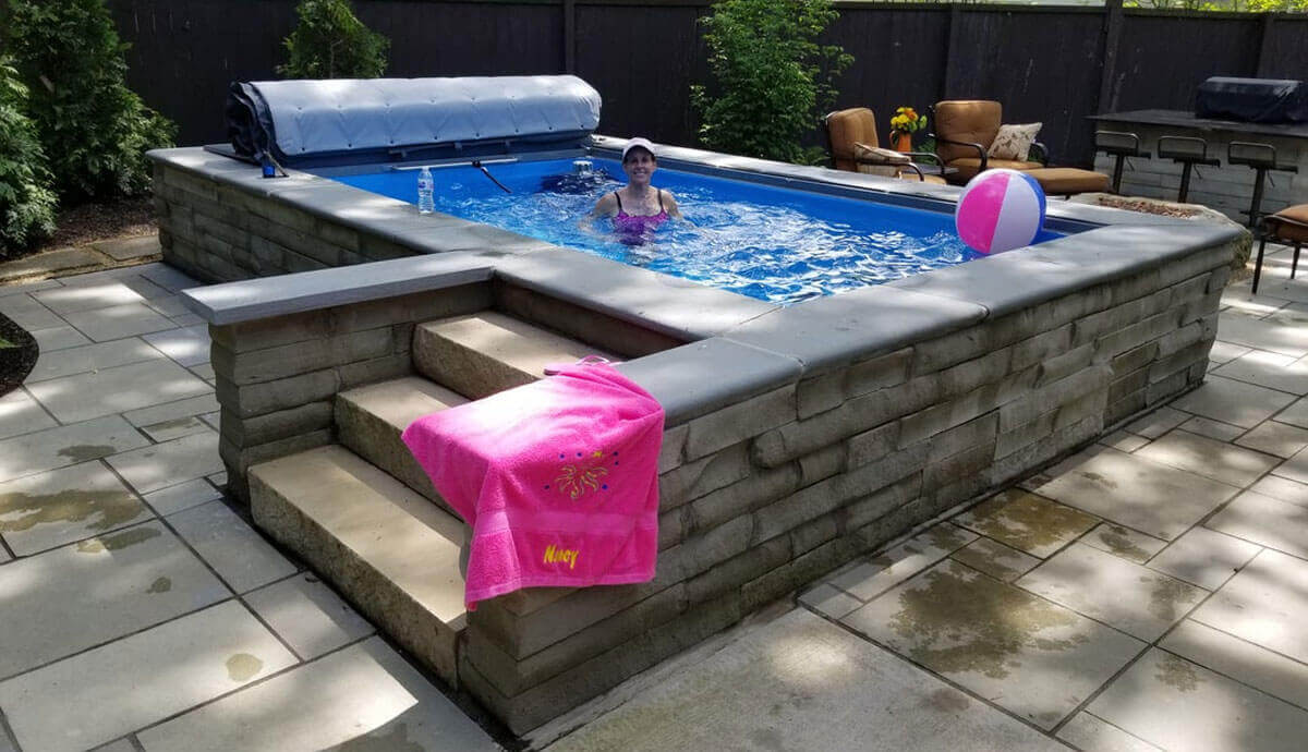 Outdoor installation of the Endless Pools Plunge Prime Therapy Pool, encased in a raised stone wall with a smooth grey coping edge. Three stone steps lead up to the pool’s edge, with a bright pink towel embroidered with the name “Nancy” draped over the top step. Inside the pool, a person is swimming toward the far end, with the white slatted cover roller in the open position. Patio seating, a barbecue, and a beach ball float are visible around the paved poolside area.
