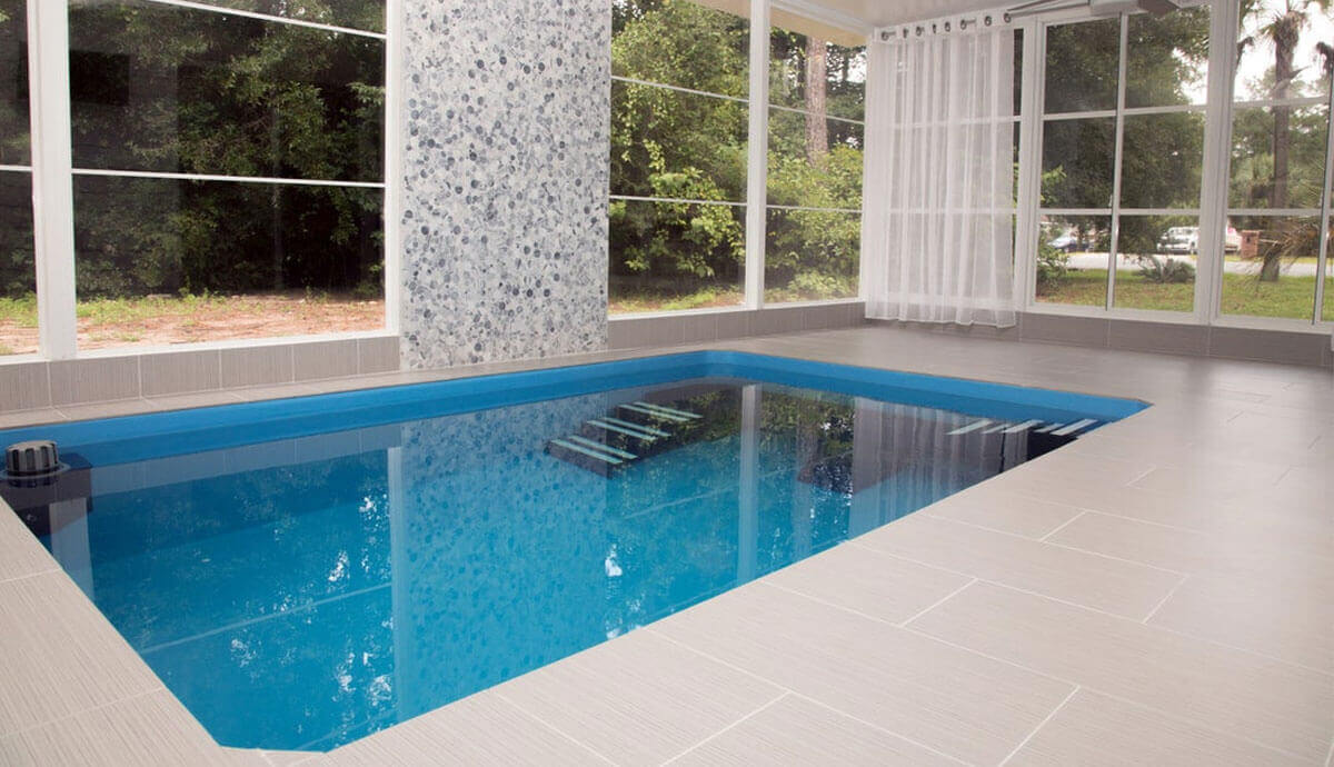 Indoor Endless Pools Plunge Prime Therapy Pool installed in a bright, tiled sunroom with full-height screened windows. The blue water reflects the surrounding trees outside. At the far wall, a decorative feature with grey and white pebble-pattern tiles extends from floor to ceiling. A slatted white pool cover roller is tucked to the left corner. The integrated bench seating is faintly visible under the calm surface.