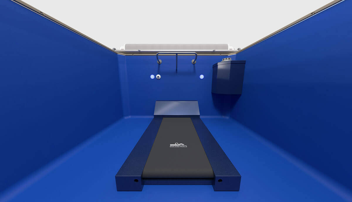 Interior view from inside the Endless Pools Plunge Prime Therapy Pool, facing towards the far wall. The bright blue walls and floor create a clean, modern appearance. Two underwater LED lights are mounted at equal heights on the far wall, with a stainless-steel grab rail positioned centrally above them. To the right side of the far wall, a grey mechanical housing unit contains the pool’s water flow and filtration system. The beige coping and partially visible slatted pool cover frame the upper edge.