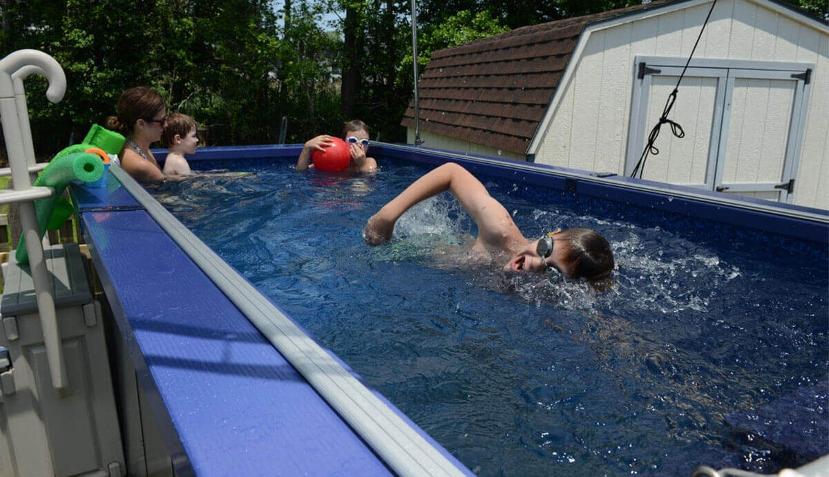 Outdoor scene of the Endless Pools SP105 Modular Pool in use. A young boy swims freestyle in the foreground, wearing swimming goggles and facing the propulsion unit. In the background, a woman and two children are at the far end, one child holding a bright red ball. The pool’s dark navy-blue coping contrasts with the shimmering blue water. A white ladder is visible on the left edge, with a green foam pool noodle hooked over it. Behind the pool, a light beige shed with double doors sits against a backdrop of green trees.