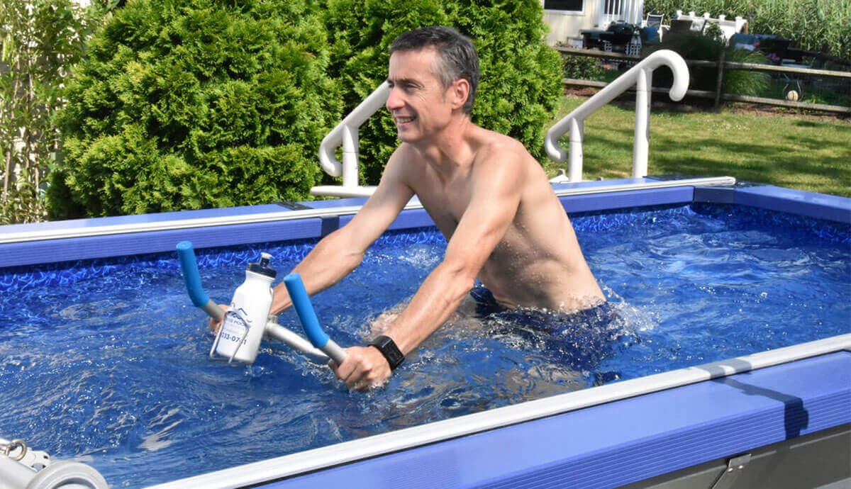 Outdoor scene of a man using an underwater stationary bike inside the Endless Pools SP105 Modular Pool. The man is positioned in the centre of the pool, holding blue handlebars with a mounted white water bottle, and smiling while pedalling through the water. The bright blue patterned liner and gentle ripples are visible around him. Behind the pool, lush green bushes and a white railing suggest a private backyard setting.