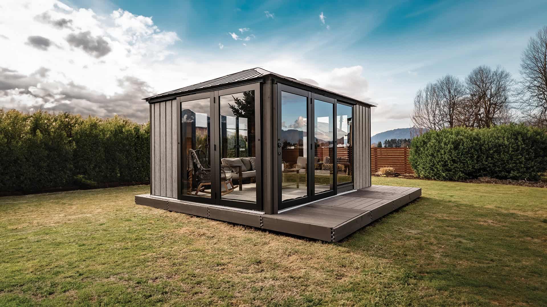 Outdoor view of the Sofia Gazebo set on a grassy lawn, angled from the front-right corner. The gazebo features black-framed glass sliding doors along the two visible sides, with wood-paneled sections at the rear. Inside, wicker and cushioned seating furniture is visible. Reflections of trees and the sky appear on the glass panels.