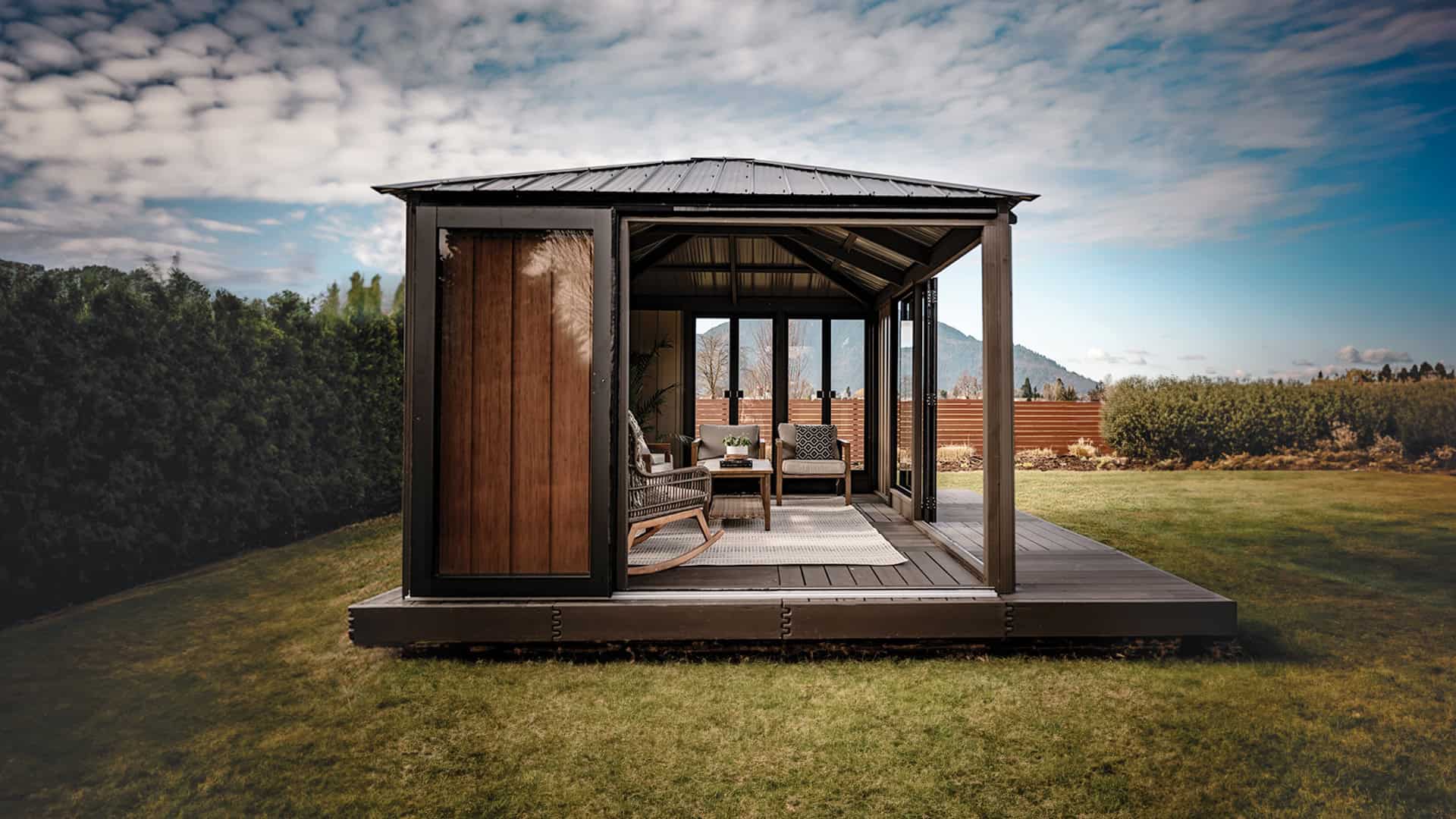 Straight-on front view of the Sofia Gazebo in a backyard setting. The structure has a black steel roof, vertical wood paneling on the left wall, and wide glass folding doors opening into the interior. Inside, wicker furniture with cushions and a central coffee table are arranged on a light rug. Behind the gazebo is a tall hedge and wooden fencing with mountains visible in the distance.