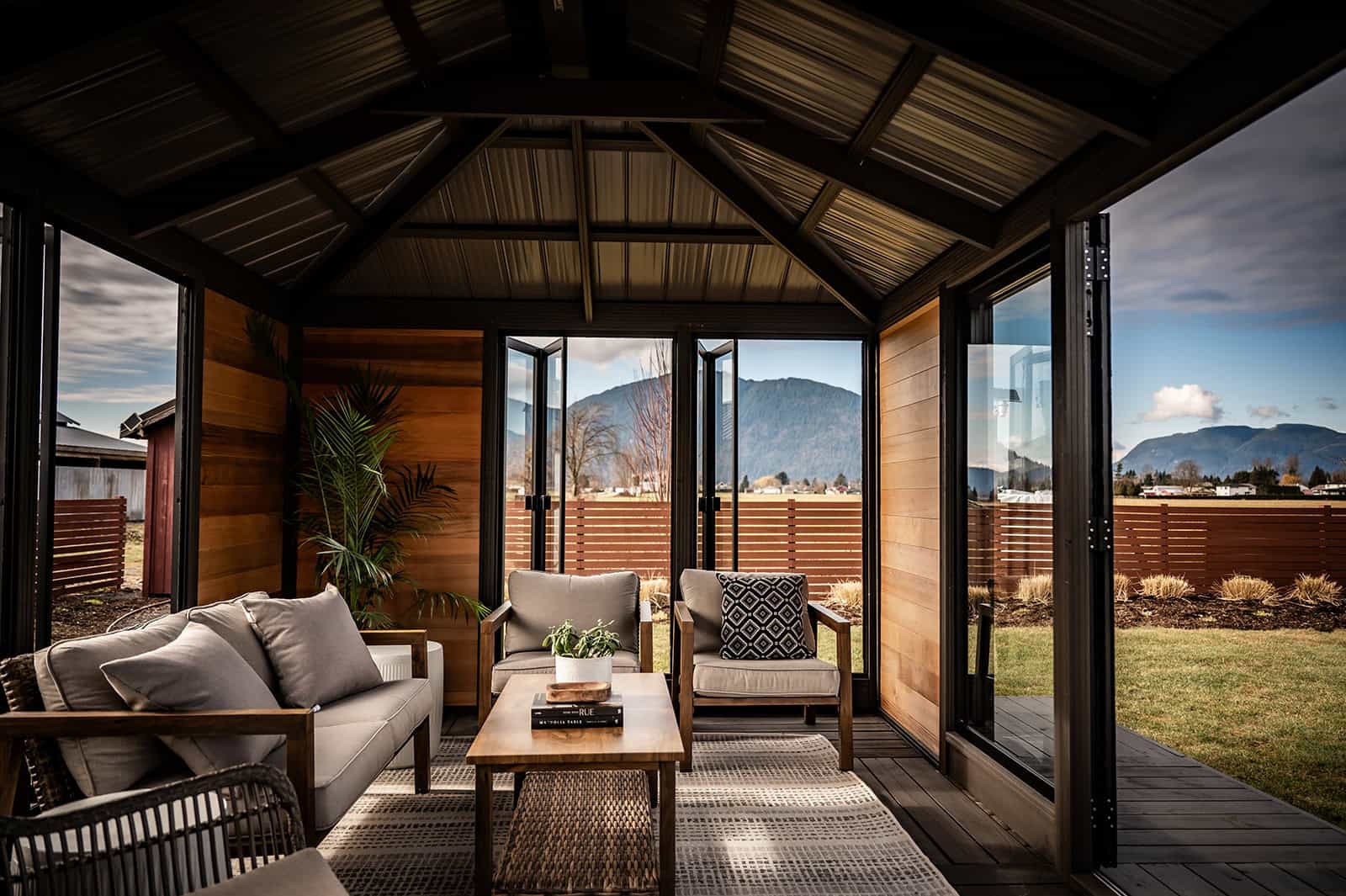 Interior view of the Sofia Gazebo facing outward through the open double glass doors. The inside walls are clad with horizontal wood panels, and the roof has exposed black steel beams. A seating arrangement with cushioned chairs and sofas surrounds a wooden coffee table on a patterned rug. Through the open doors, a wooden fence, grassy yard, and mountain range are visible.