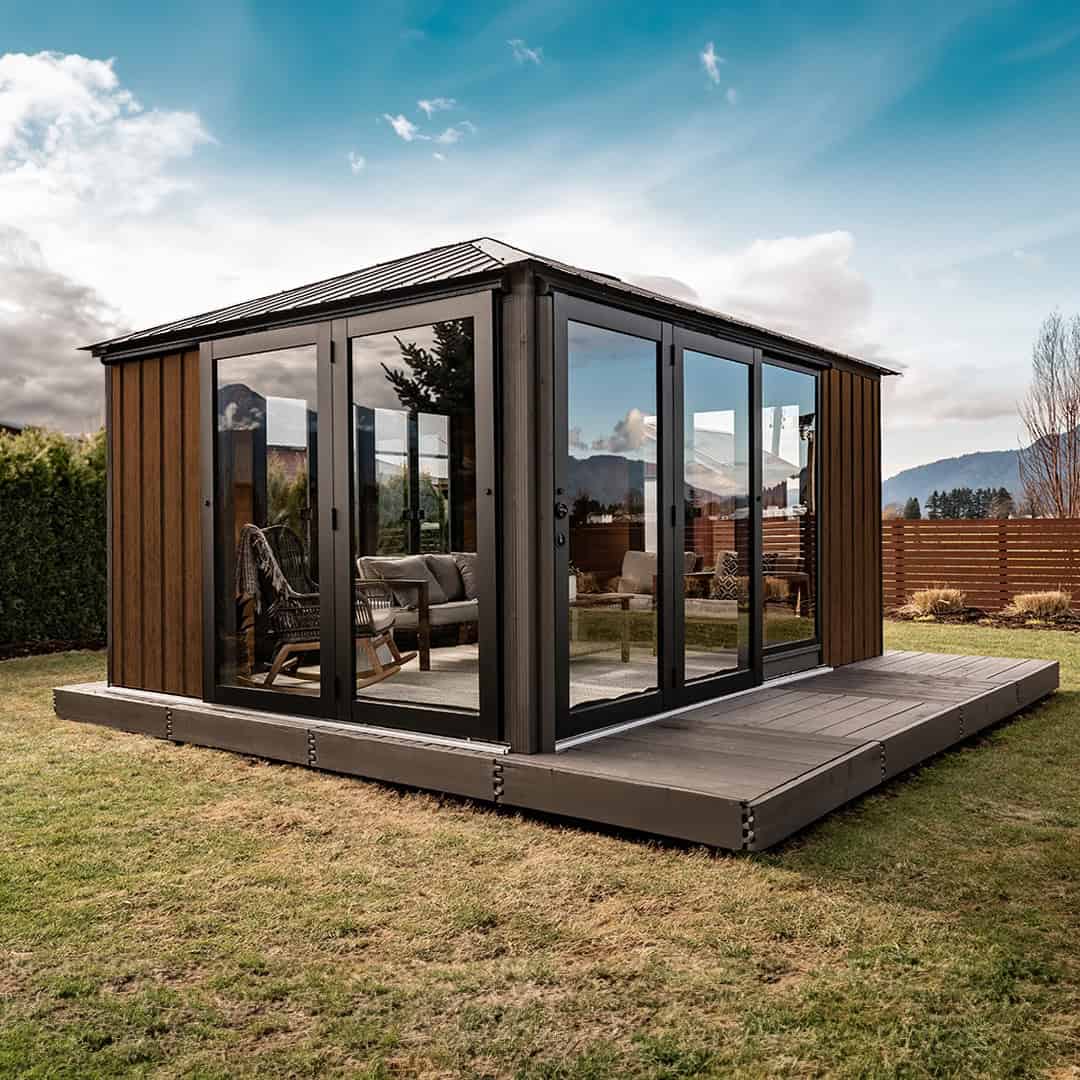 Exterior corner view of the Sofia Gazebo on a lawn, seen from the left-front angle. The gazebo features glass sliding doors with black frames across two sides, and wood-paneled wall sections on the far side. Inside, outdoor lounge seating with cushions is visible. The backdrop includes hedges, bare trees, and distant mountains beneath a partly cloudy sky.