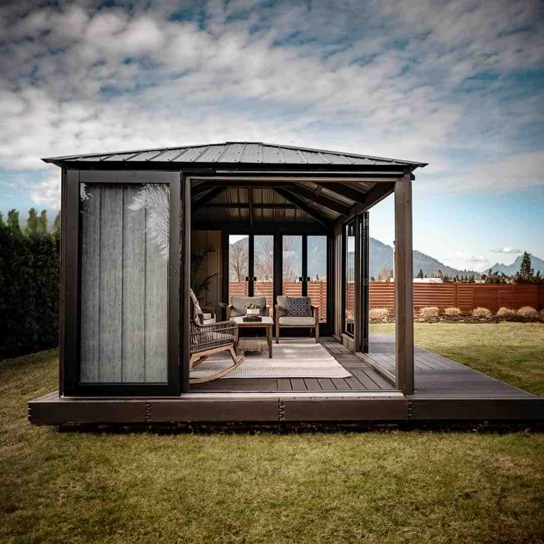 Frontal outdoor view of the Sofia Gazebo with wide black-framed folding glass doors partially open, revealing cushioned seating inside. The left side panel is solid wood, while the right side has glass. The roof is pitched steel with dark ridges. The gazebo sits on a raised wooden base, with mountains and hedges in the background.