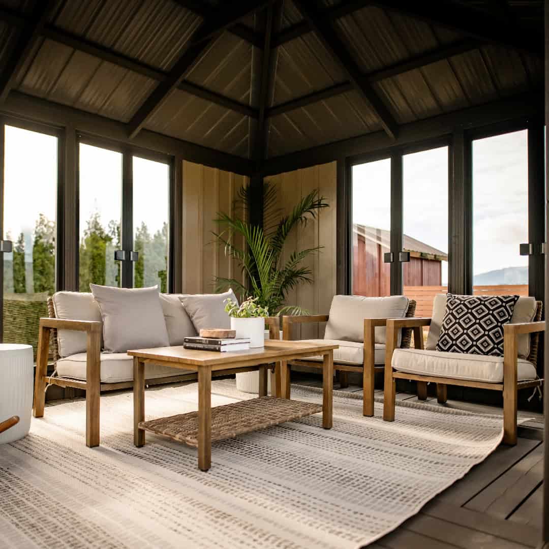 Close-up view of the Sofia Gazebo interior, showing a lounge arrangement with light beige cushioned chairs and sofa around a wooden coffee table placed on a patterned beige rug. A potted green plant stands in the rear corner, next to black-framed glass doors. Sunlight illuminates the space, highlighting the wood paneling and warm, cozy atmosphere.