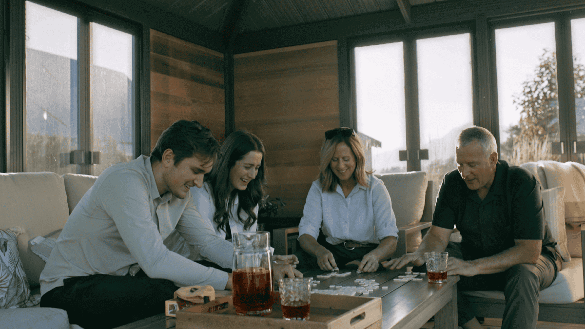 Indoor lifestyle render of the Sofia Gazebo with four adults seated around a wooden coffee table, playing a board game. Two women and two men are smiling and leaning forward, with drinks placed on the table. The background shows wooden paneled walls, large glass windows, and partial outdoor scenery.