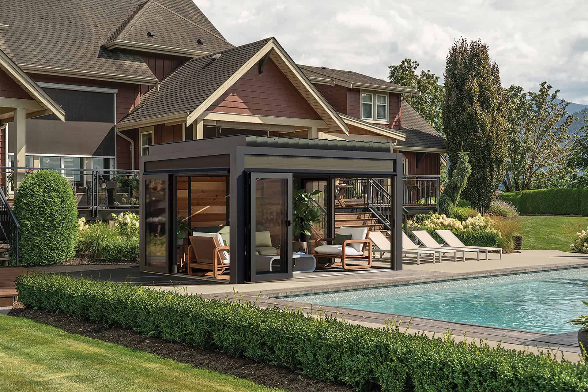 Outdoor installation of the Stella Fully Enclosed Pergola positioned beside a swimming pool in a landscaped garden. The pergola features a black powder-coated aluminium frame with transparent sliding glass doors and warm wood interior wall panels. Inside, cushioned wooden lounge chairs are neatly placed against the back wall. Decorative greenery surrounds the pergola, while a large modern house and additional seating are visible in the background.