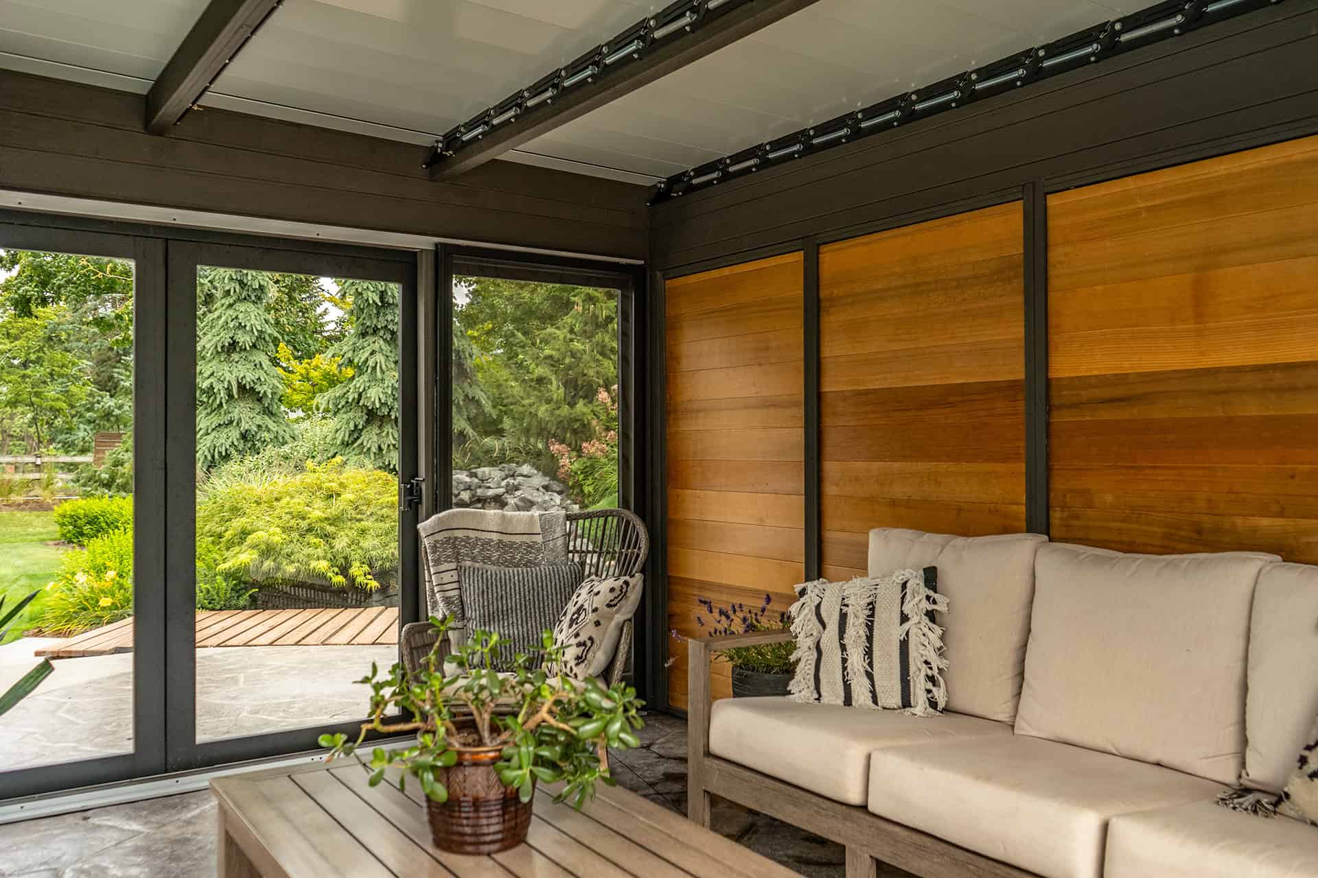 Interior detail of Stella Fully Enclosed Pergola, showing a right wall made of vertical wooden panels and left wall composed of large black-framed glass sliding doors. A beige sofa with multiple patterned pillows lines the wood wall, complemented by a low wooden coffee table with a green plant in a ceramic pot. Outside through the glass, dense garden greenery is visible.